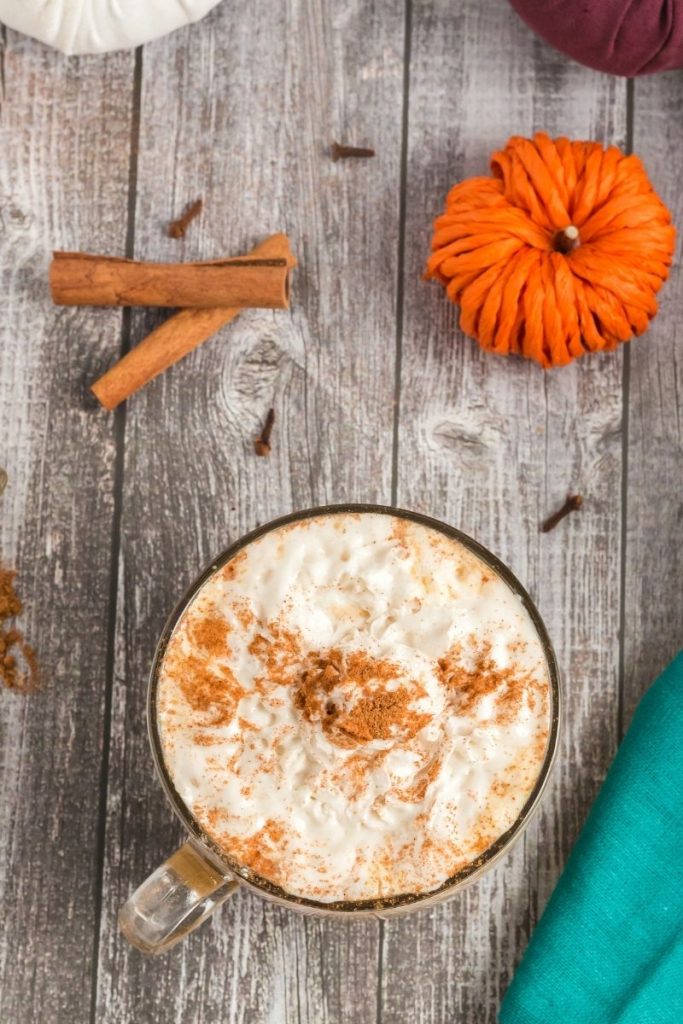 top view of a pumpkin spice latte in a large round glass mug. You can only see teh whipped cream and sprinkled cinnamon. Around the mug are cinnamon sticks and an orange pumpkin