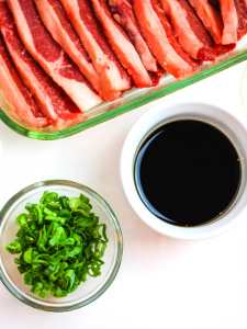 a glass pan of short ribs, a round bowl of soy sauce and green onions