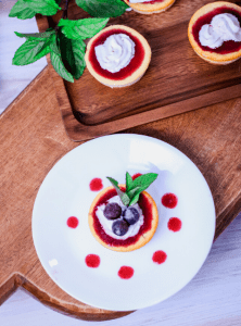 a top view of a mini cheesecake on a whilte plate decorated with whipped cream, blueberries and a mint leaf