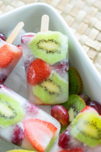 top view of popsicles in a white bowl of ice. The popsicles have kiwi and strawberry slices and grapes