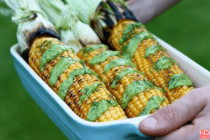 grilled corn in a white serving dish with green dressing drizzled over it