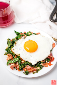 a fried egg on top of a plate of prosciutto and kale with a red drink and the handle of the cast iron pot in the corner