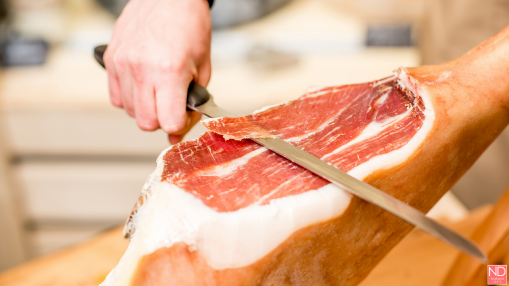 a hand thinly slicing prosciutto from a dried pork leg
