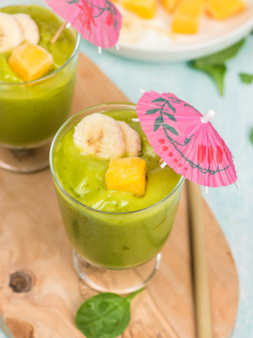 45 degree view of a green smoothie with slices of bananas, a square of mango and a pink unbrella