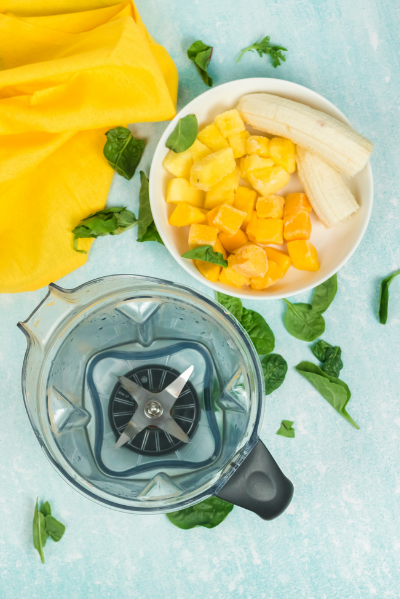 top view of a vitamix container with coconut water and a white bowl on the side with a banana, frozen mango and frozen pineapple. spinach leaves are sprinkled around the picture and there is a yellow napkin in the top left corner