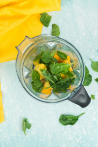 top view of a viatmix blender container with frozen mango and pineapples and fresh spinach leaves. The container is on a light blue background with a yellow towel in the top left corner