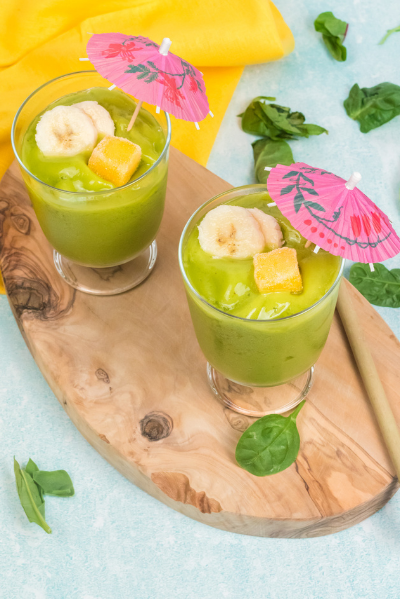 two green smoothies on a oval wooden cutting board with spinach leaves sprinkled.