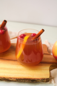 a side view of a stemless wine glass with cloudy apple juice and garnished iwtwh an orange slice and cinnamon stick