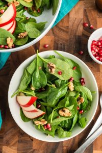 top view of a simple pear salad with pomegranates and walnuts white bowl with spinach leaves, pomegranates, sliced red pears and walnuts. The larger bowl is peeking in in the top left hand corner