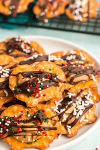 45 degree view of a white plate with pretzel bites on them. Each pretzel bite is a flat pretzel crisp with a ball of peanut butter drizzled with melted chocolate and topped with red, green and white sprinkles