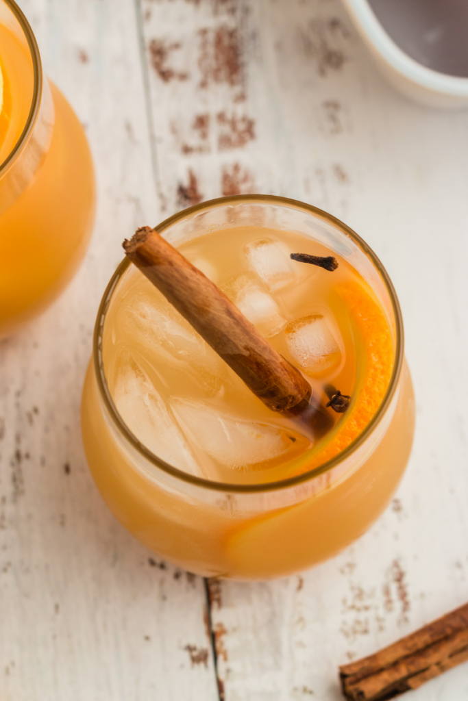 top view of a glass with apple cider. It has ice, a cinnamon stick, a round orange slice and whole cloves on top