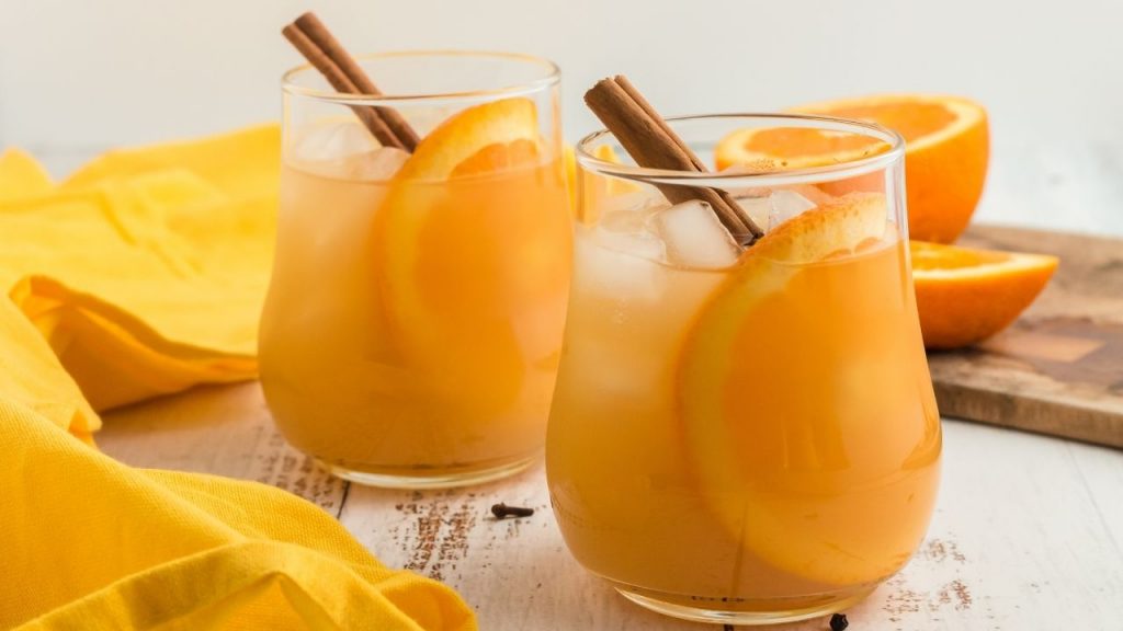 two glasses of apple cider each with a round orange slice showing on through the glass and a cinnamon stick popping out the top
