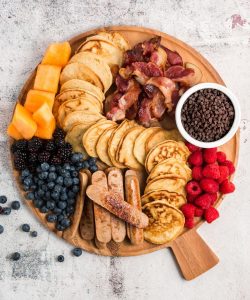 top view of a round board with pancakes down the middle, fruits, sausage and bacon