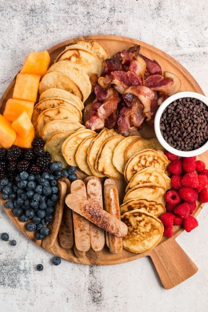 top view of round wooden board with pancakes, fruits, sausage and bacon
