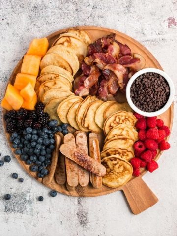top view of a round board with pancakes down the middle, fruits, sausage and bacon
