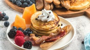 a stack of pancakes with whipped cream and chocolate chips on tap and bacon, sausage and fruits on the side