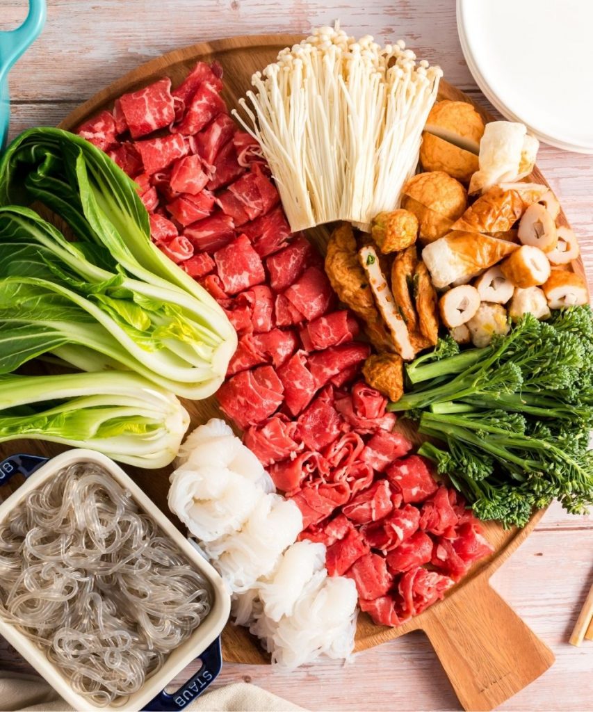 top view if a round wooden tray with meat, baby bok choy, broccoil, mushrooms, fish cake and noodles
