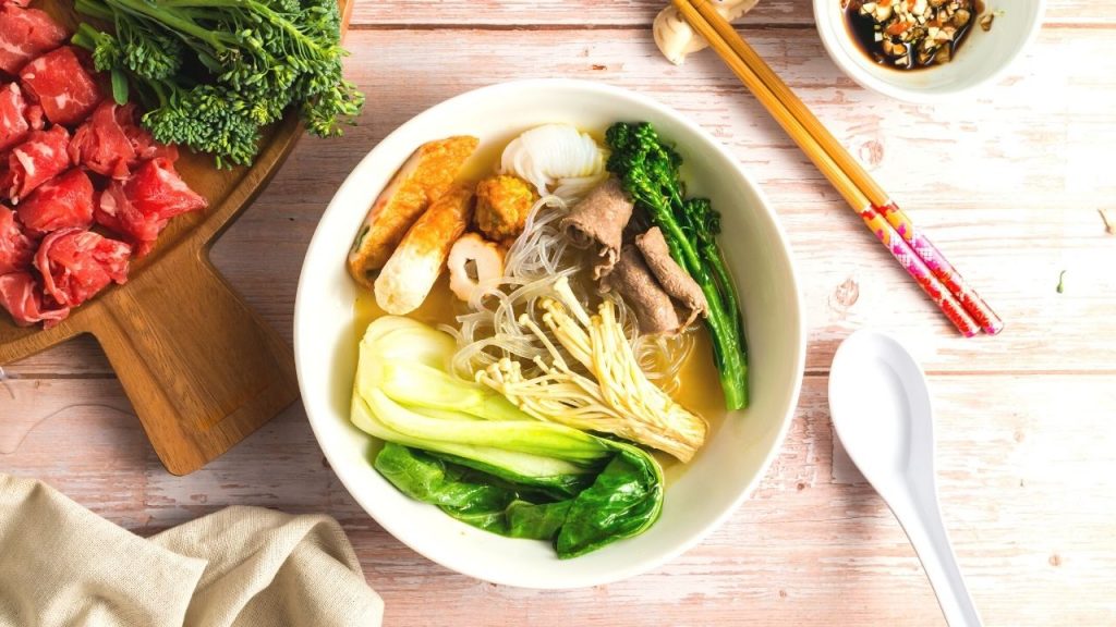 top view of a bowl of broth, mushrooms, baby bok choy, fish cake, beef and broccoli