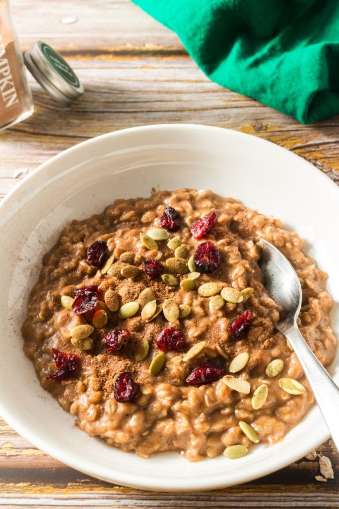side view of a white bowl with cooked oatmeal topped with cranberries and pumpkin seeks with a spoon in the bowl