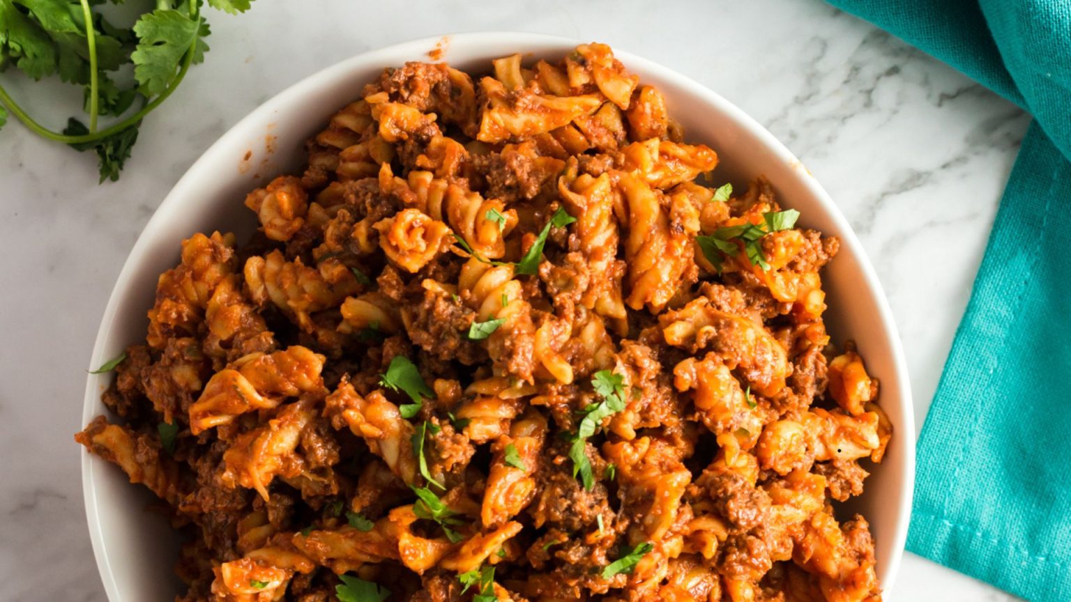 A bowl filled with rotini pasta mixed with a chunky tomato sauce and ground beef garnished with fresh parsley. The dish is placed on a marble surface with a sprig of cilantro and a turquoise cloth nearby.